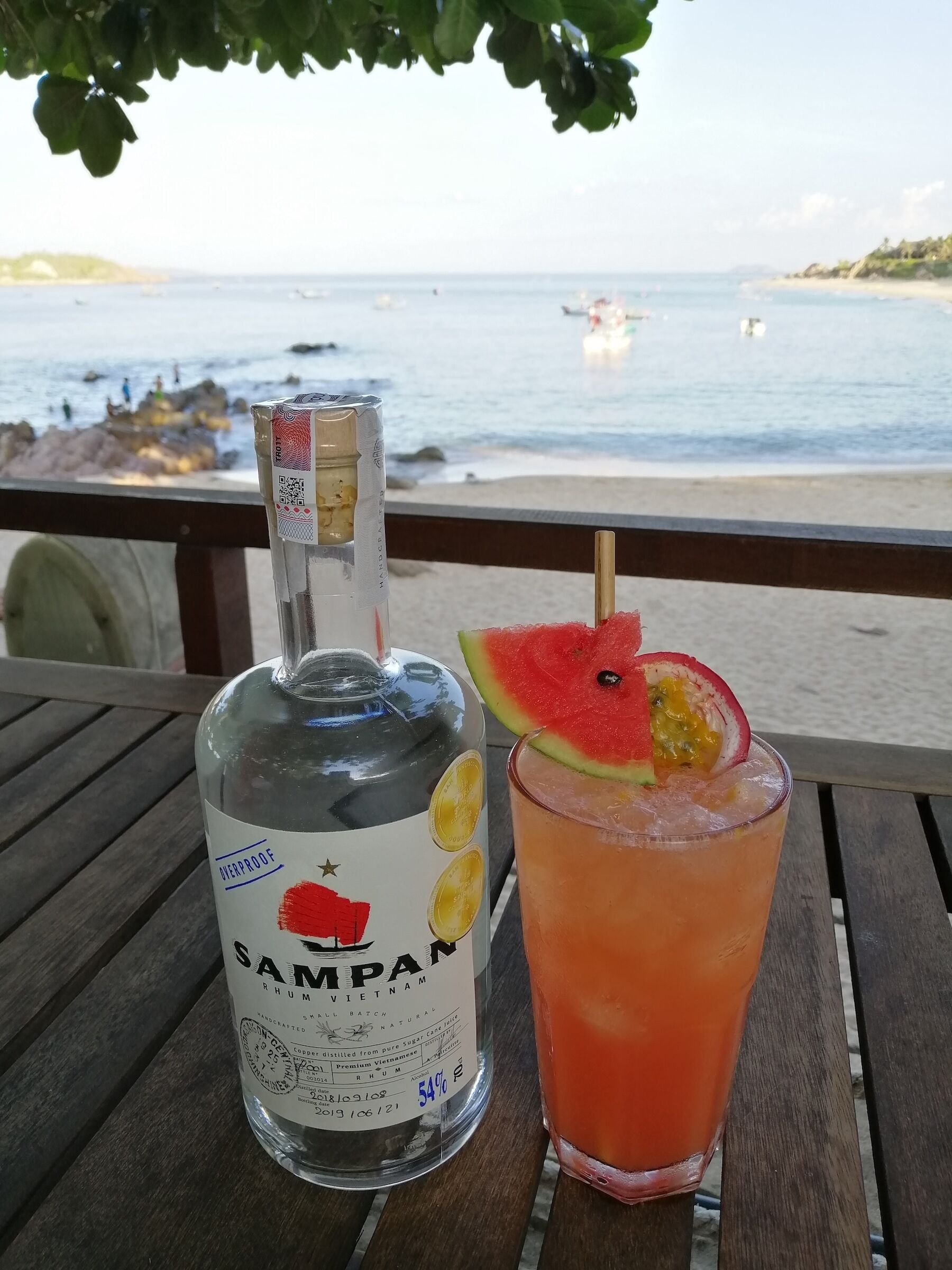 A bottle of Sampon rum and a glass of orange drink with a watermelon slice on a wooden table by the beach.