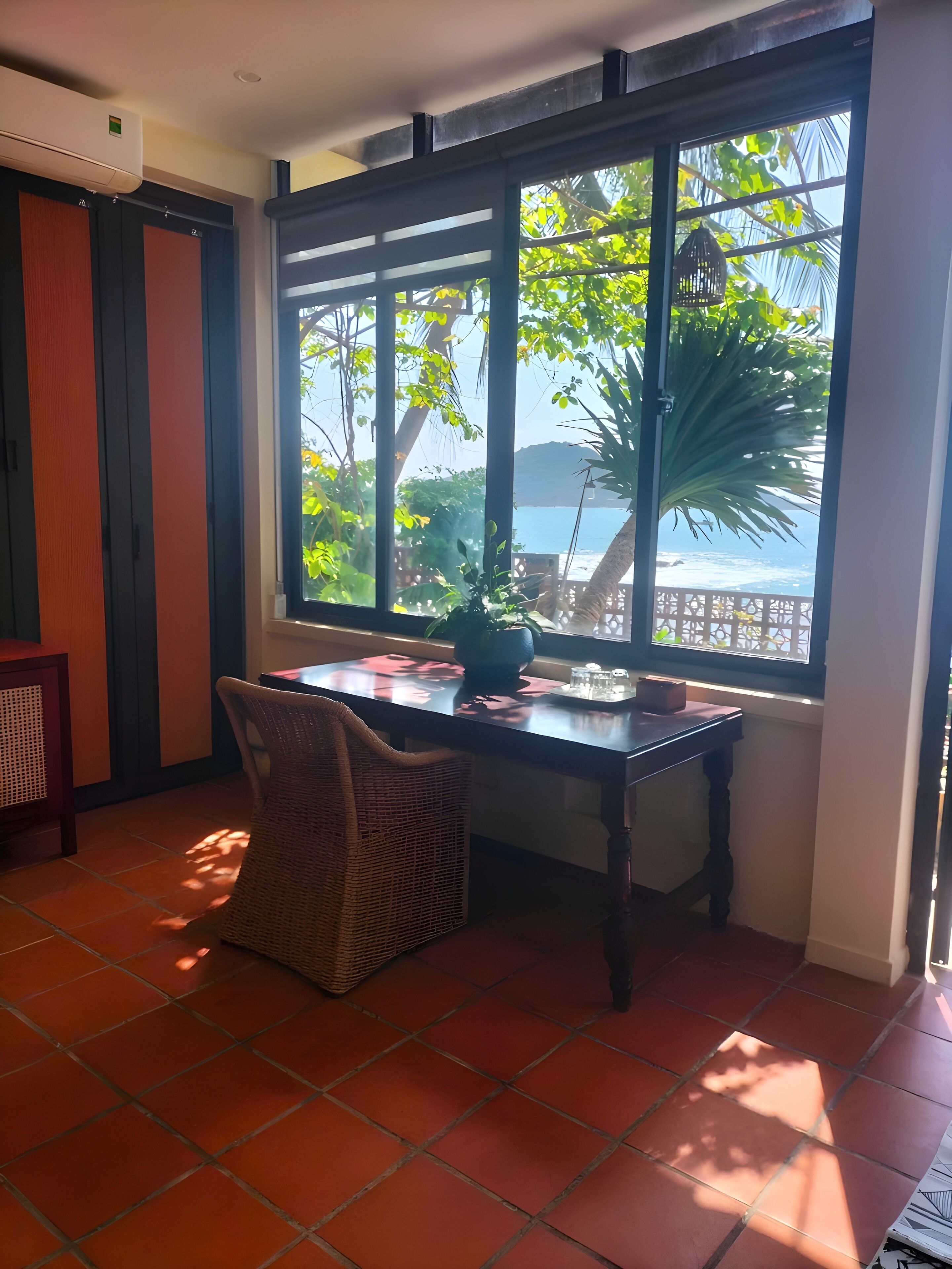 A room with a large window, a table, a wicker chair, and a tiled floor.