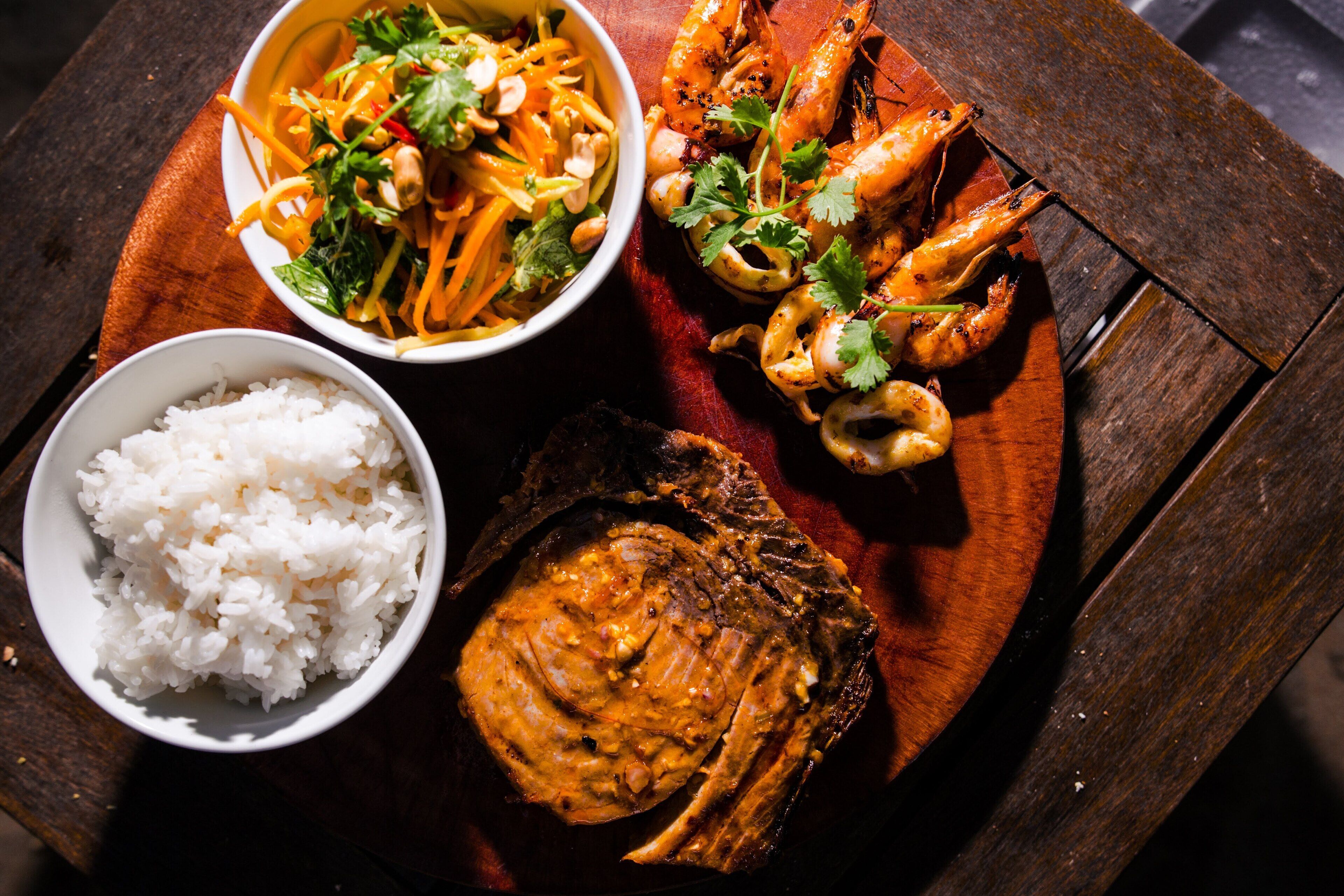 A wooden table with a bowl of white rice, a bowl of stir-fried vegetables, grilled shrimp, and a piece of grilled meat.