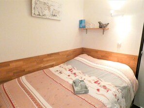 A bedroom with a wooden headboard, a bed with a patterned bedspread, a bedside table with a cup holder, and a wall-mounted shelf with decorative items.