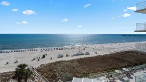 A beach with a pier in the distance, a sandy beach area, and a building with a balcony overlooking the beach.