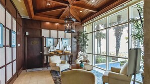 A spacious lobby with a high ceiling, wooden beams, and large windows offering a view of palm trees. There are several seating areas with chairs and a small table, and a reception desk with framed pictures and a sign.