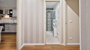 Un couloir menant à une salle de bain avec douche, un coin cuisine équipé de placards et d’un plan de travail, et un plancher en bois.