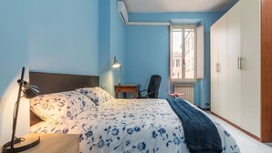 Un dormitorio con cama, escritorio, silla, armario y ventana con vistas a edificios.