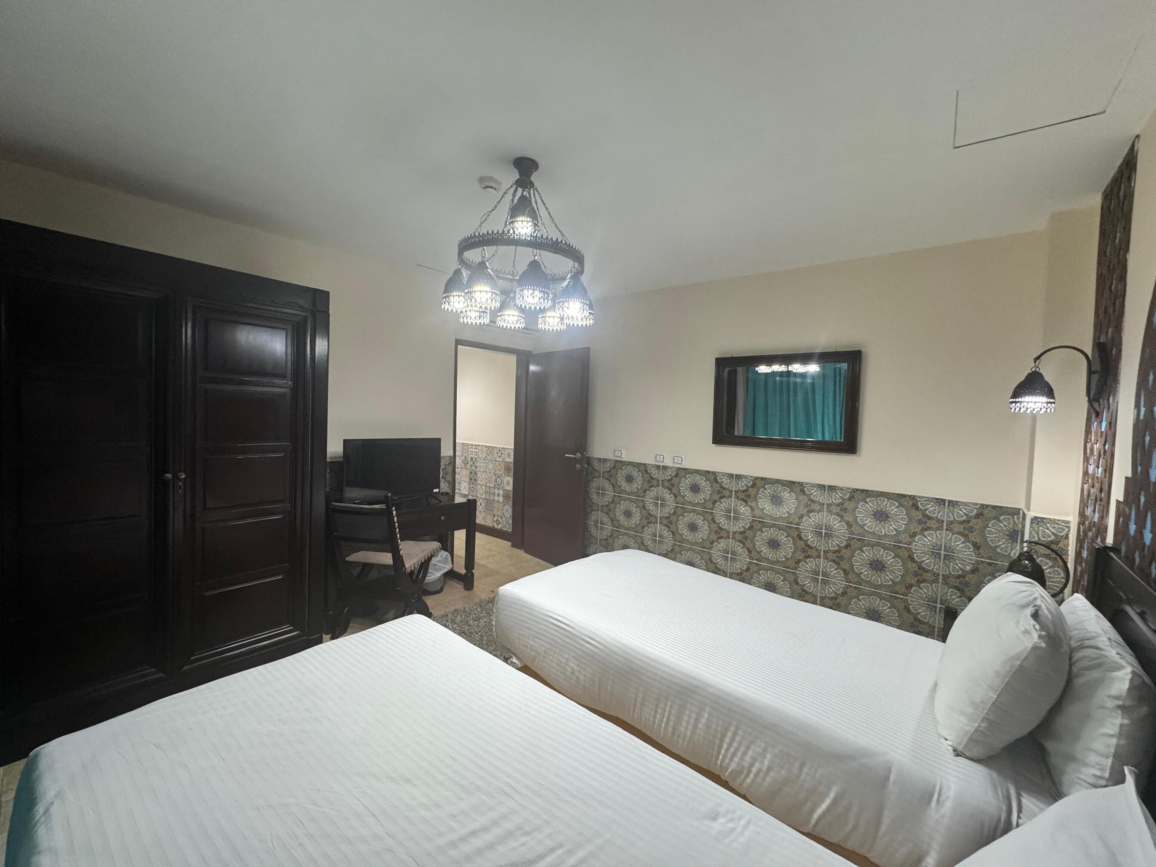 Habitación de hotel con dos camas, un escritorio, una silla, una araña y azulejos con diseño en la pared.