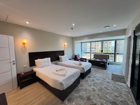 A hotel room with two beds, a desk, a chair, and a large window offering a city view.