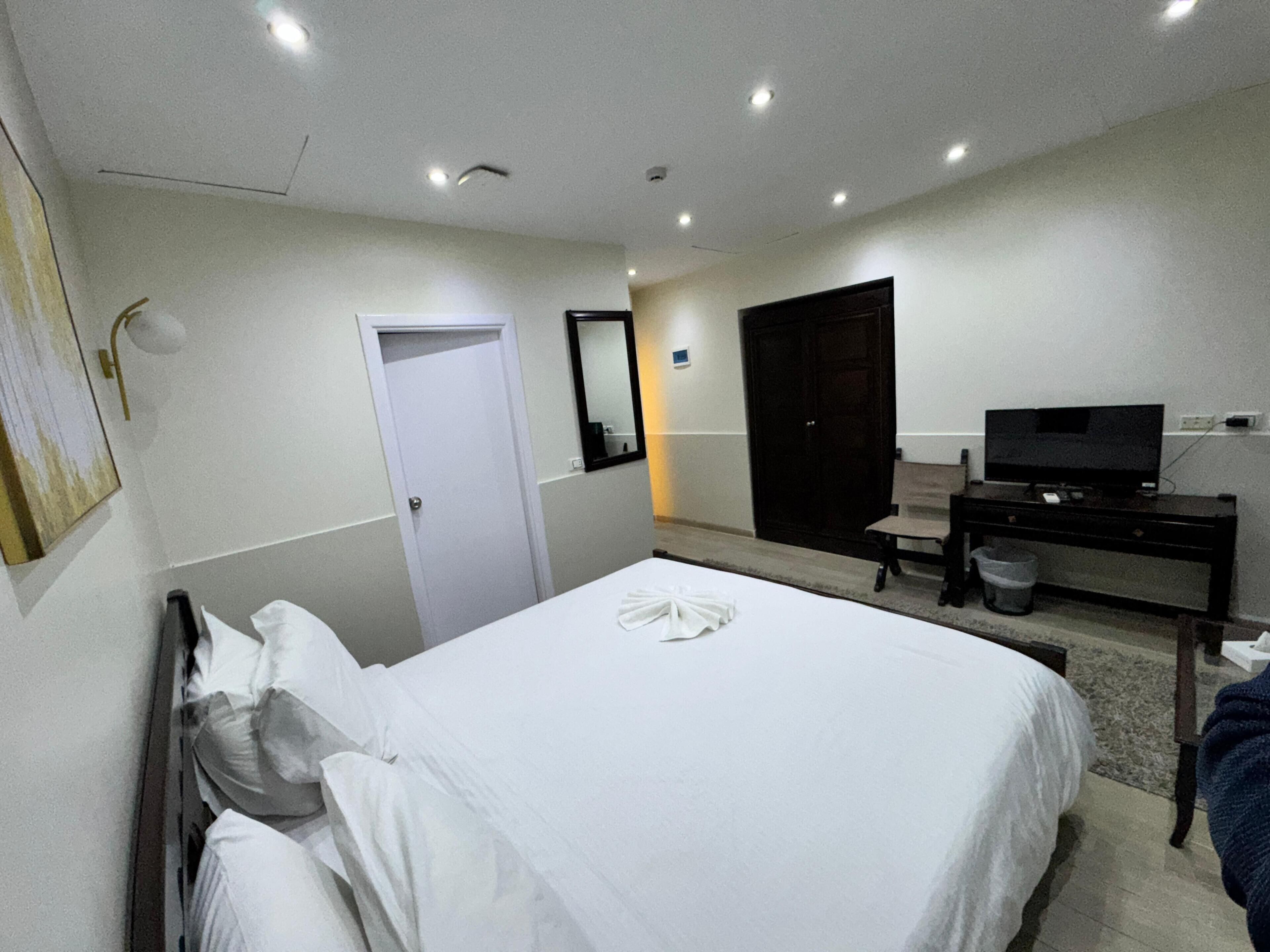 A hotel room with a bed, a desk with a television, a door, and a wall-mounted mirror.