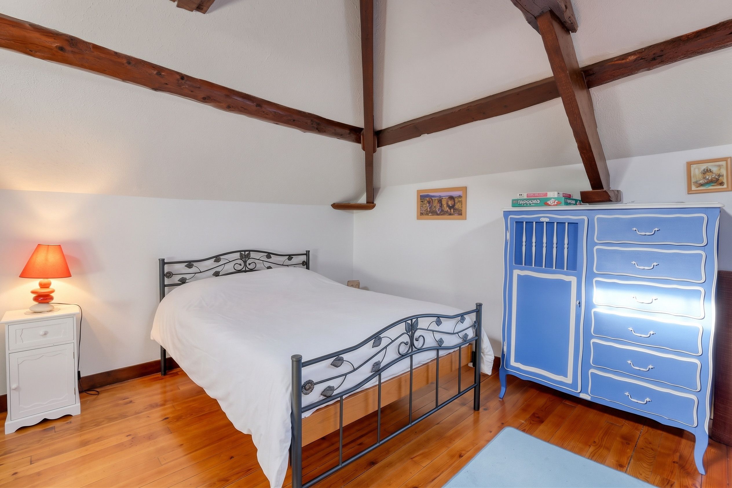 A bedroom with a single bed, a nightstand with a lamp, a dresser, and wooden beams on the ceiling.