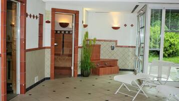 Um saguão de hotel com piso de azulejos, balcão de recepção, área de estar com cadeiras brancas e uma grande planta em vaso.