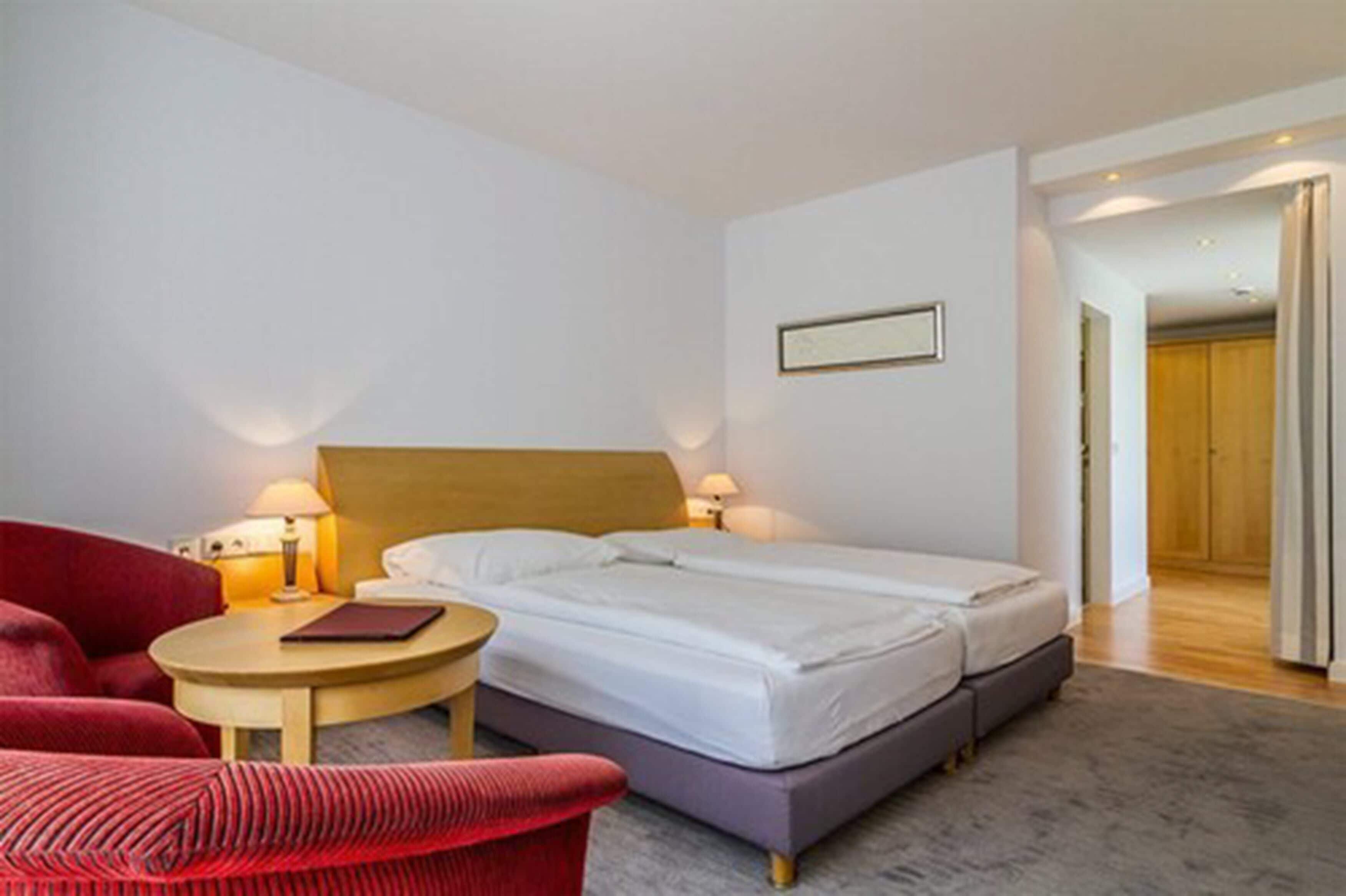 Una habitación de hotel con una cama, un sillón rojo, una mesita redonda y una lámpara.