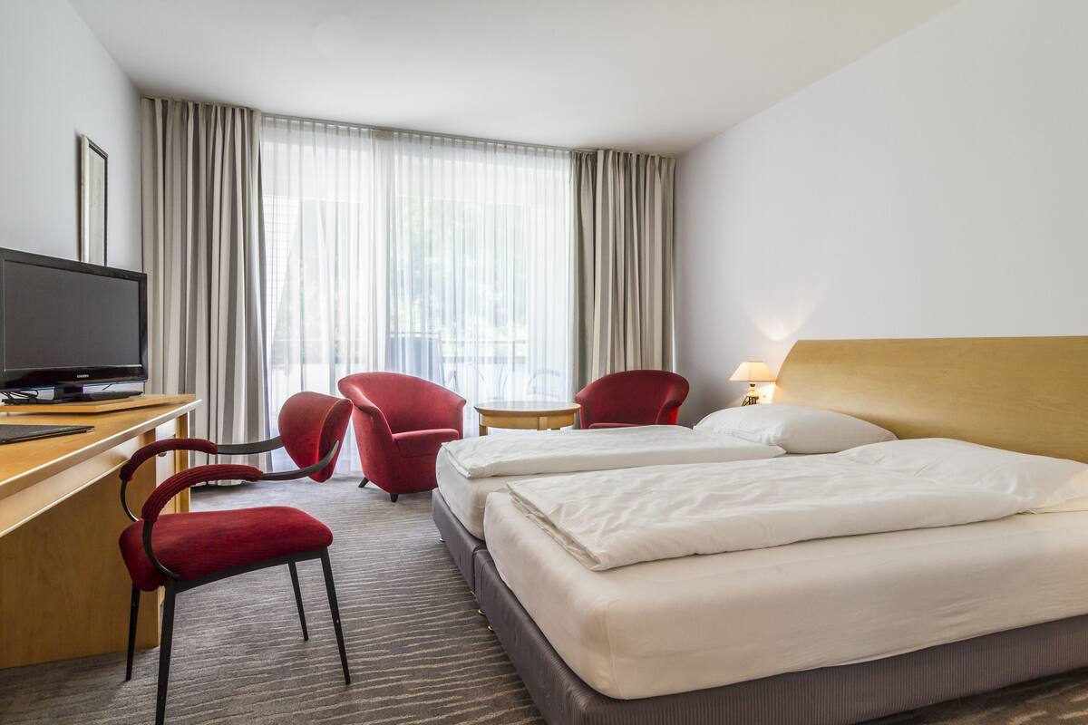 Une chambre d’hôtel avec deux lits, un bureau avec un ordinateur, deux chaises rouges, une petite table et un téléviseur.