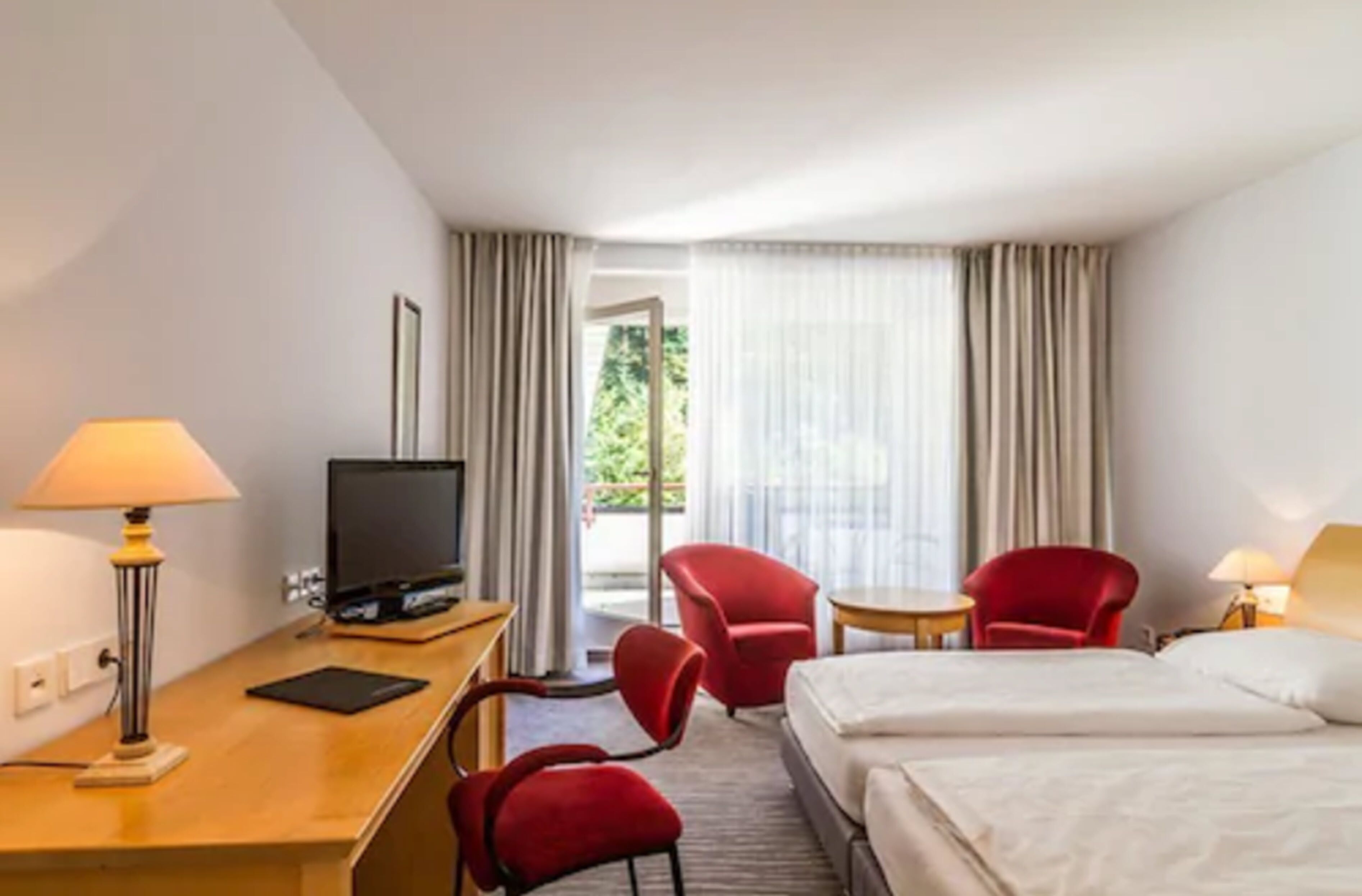 A hotel room with a bed, a desk with a computer, a TV, two red chairs, and a window with curtains.