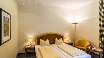 Une chambre d’hôtel avec deux lits, une table de chevet avec une lampe, une chaise et une petite table avec des fruits.
