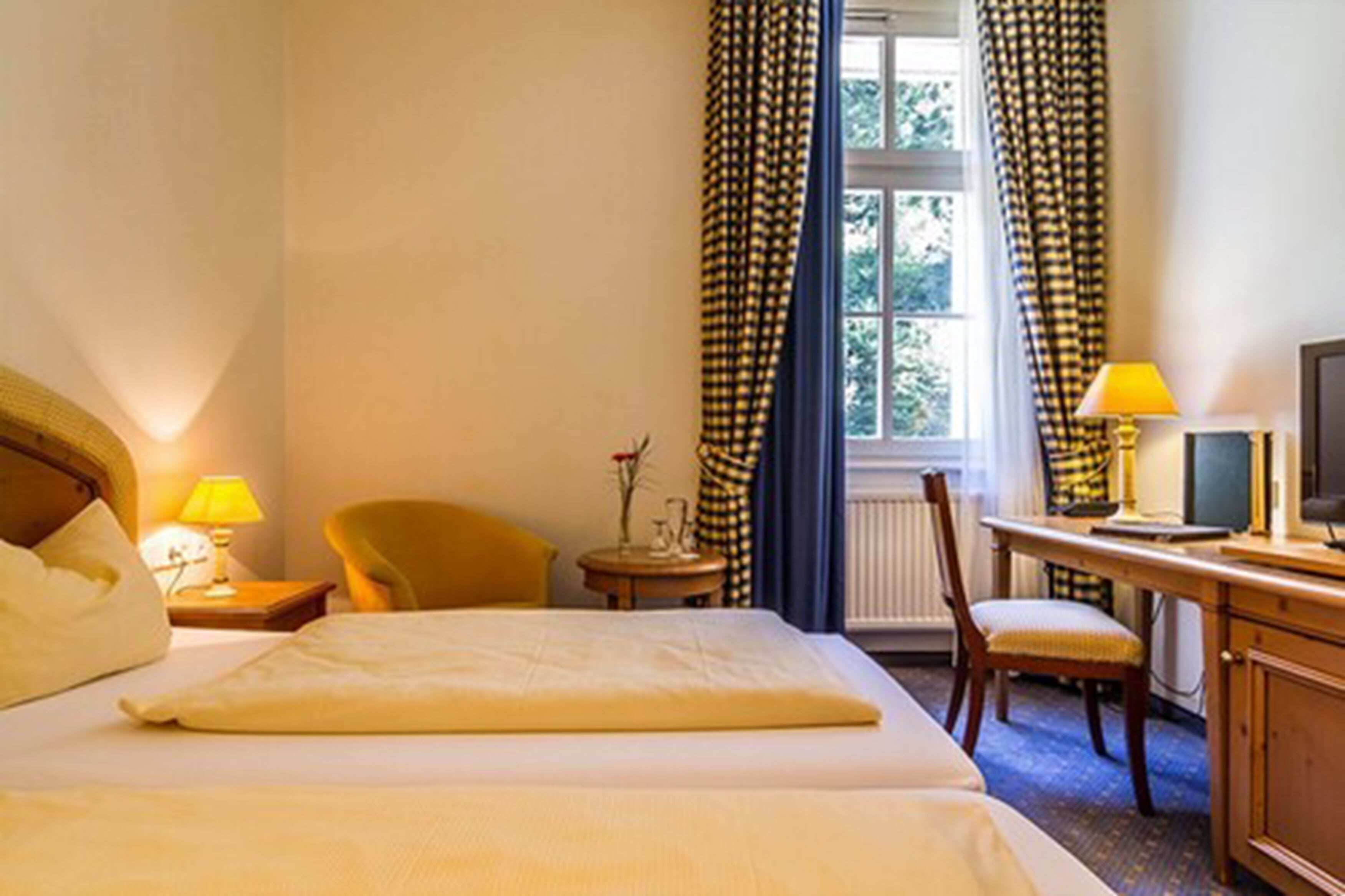 Quarto de hotel com uma cama, escrivaninha com cadeira, luminária, televisão e janela com cortinas.