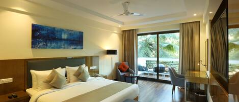 A hotel room with a large bed, a desk, a chair, a TV, and a balcony with a view of palm trees.