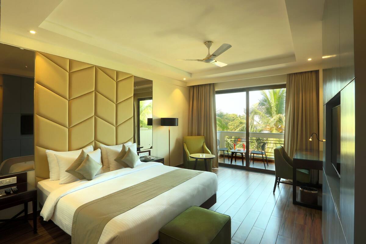 A hotel room with a large bed, a desk, a chair, a TV, and a balcony with a view of palm trees.