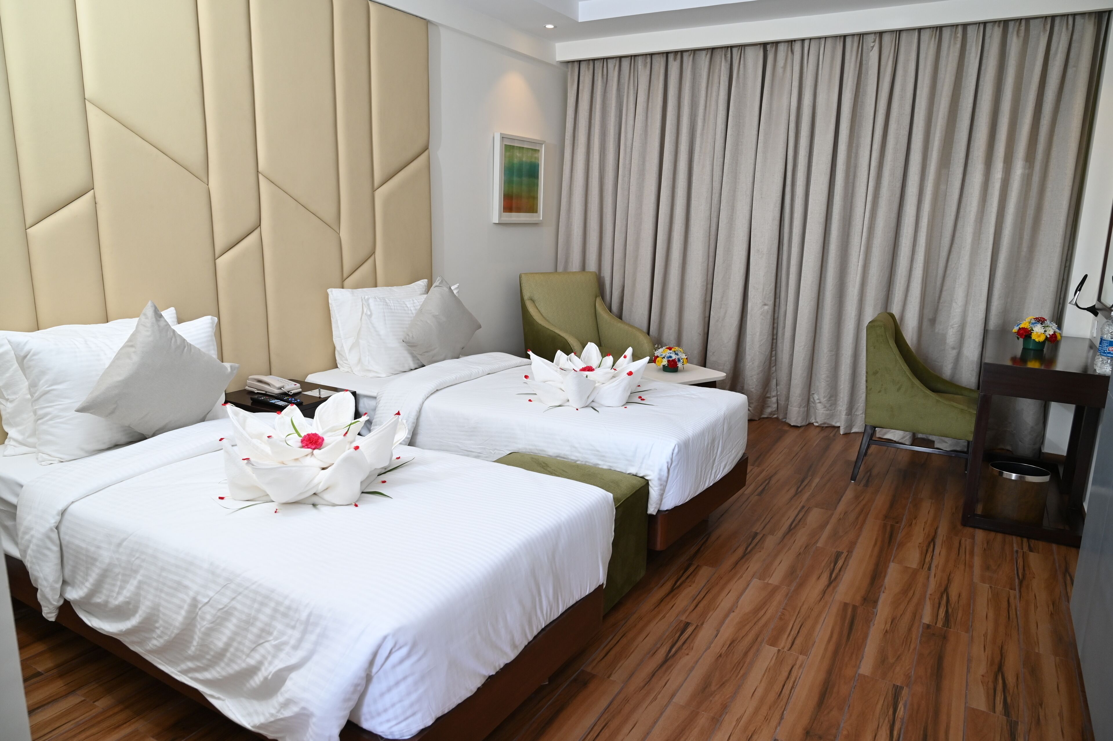 A hotel room with two beds, a green armchair, a small table with a vase of flowers, and a wooden floor.