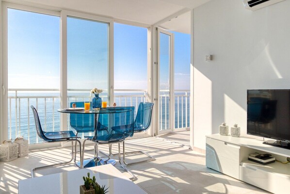A balcony with a glass table and chairs, a TV, and a view of the sea.