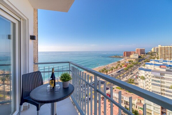 A balcony with a table, a chair, a bottle, and a potted plant, overlooking a beach and cityscape.