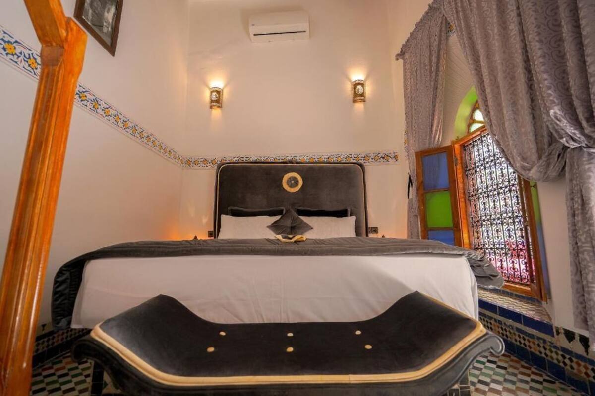 Quarto com uma cama grande, duas luminárias fixadas na parede, uma janela com grades de ferro decorativas e um piso estampado.