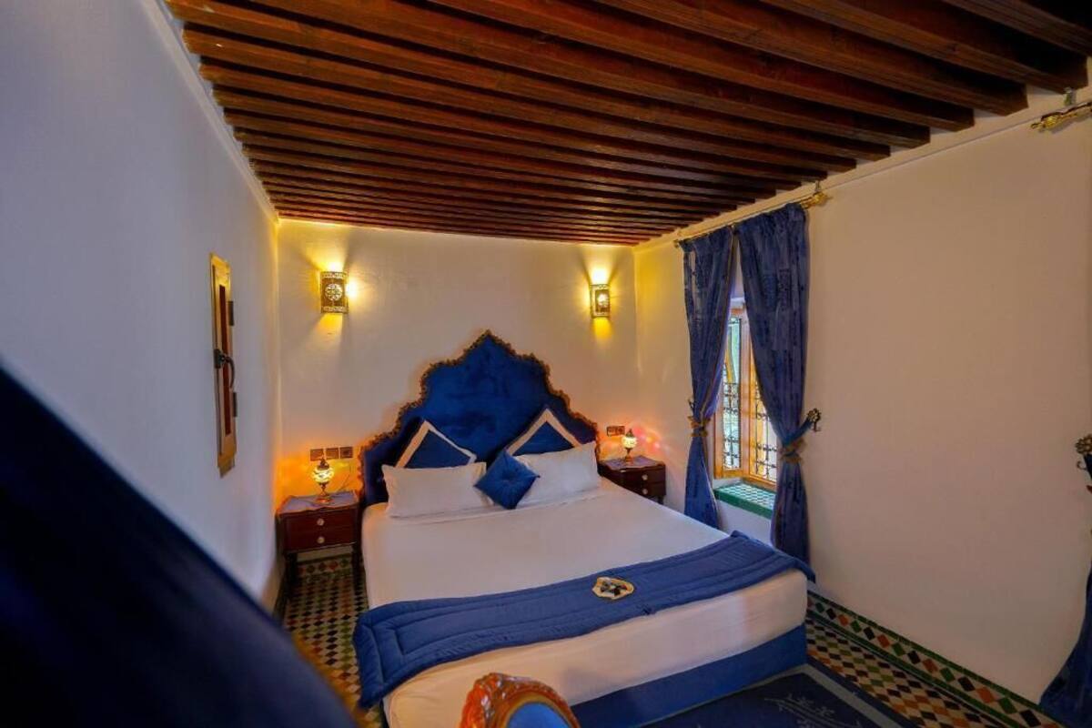 Quarto com teto de madeira, cama com cabeceira azul, duas luminárias de apoio e uma janela com cortinas.