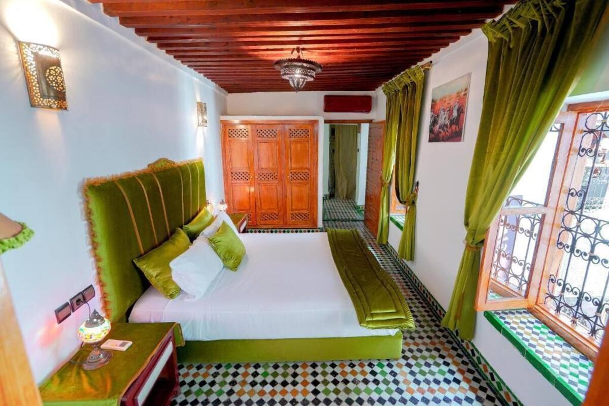 Quarto com cama, mesa de cabeceira, abajur, guarda-roupa de madeira e janela com cortinas.