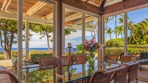 A glass-top dining table with wicker chairs, surrounded by a glass railing, overlooking a scenic view of the ocean and palm trees. - Fabulous Ocean Views with Mauna Kea Resort Access (Waimea)