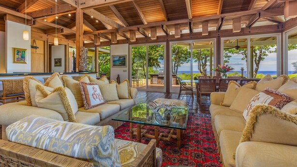 A spacious indoor area with a high ceiling and wooden beams, furnished with beige sofas and a glass coffee table, leading to an outdoor patio with a view of the ocean. - Fabulous Ocean Views with Mauna Kea Resort Access (Waimea)
