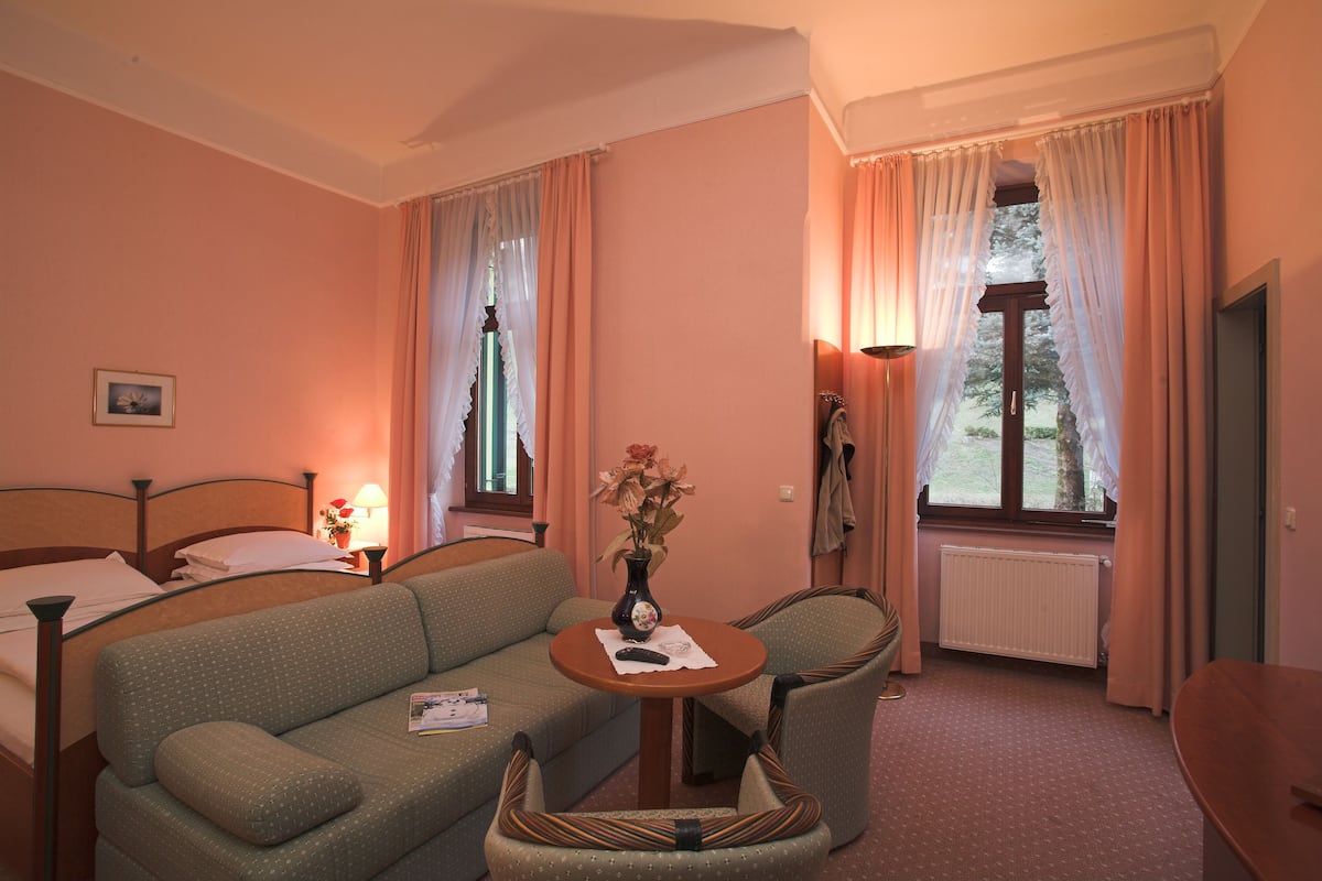 A hotel room with a bed, a sofa, a small table with a vase, and a window with curtains.