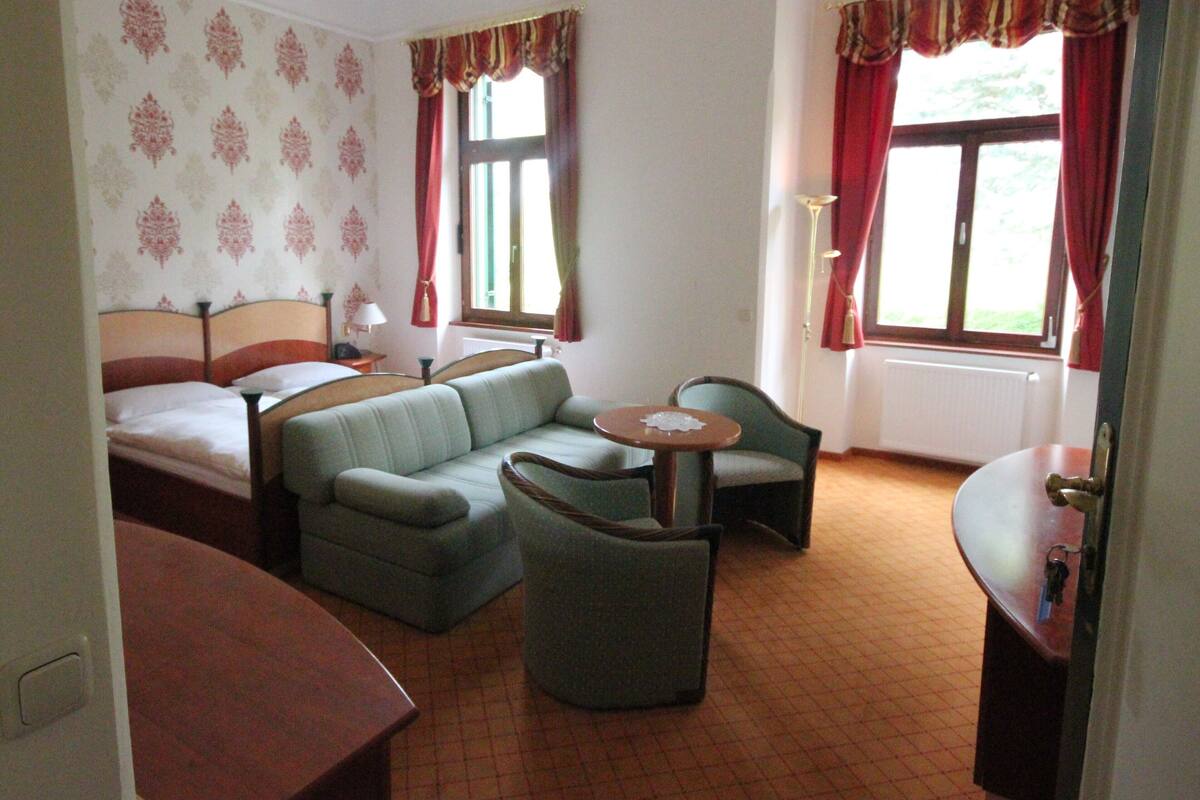 A hotel room with a bed, a sofa, a chair, a small table, and two windows with curtains.