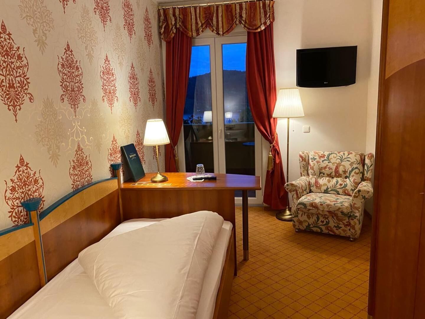 Une chambre d’hôtel avec un lit, un bureau avec une lampe, une chaise, une télévision et une fenêtre avec des rideaux.
