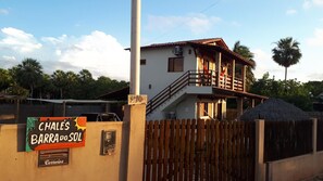 A two-story building with a balcony, a wooden fence, and a sign for 'Chales Barrado Sol'. - Chalés Barra do Sol (Cajueiro da Praia)