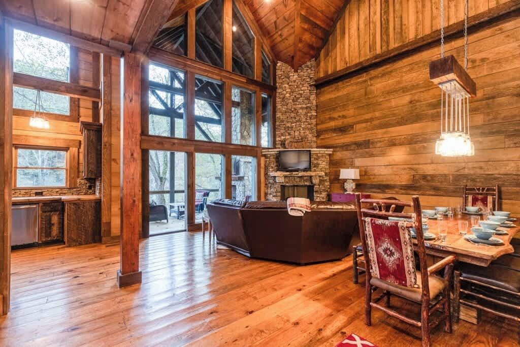 A rustic cabin interior with a stone fireplace, wooden dining table, and large windows.