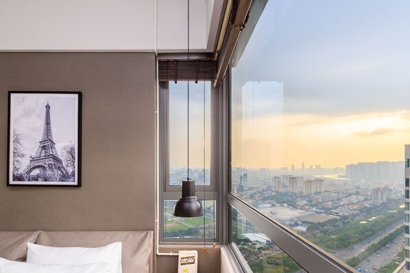A hotel room with a large window offering a view of a cityscape, a framed picture of the Eiffel Tower, and a bedside table with a lamp.