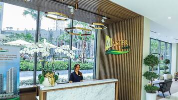 A modern hotel reception area with a wooden reception desk, a receptionist, and a view of the cityscape through large windows.