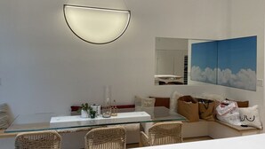 Een moderne eetruimte met een glazen tafel, rieten stoelen, een hanglamp en een spiegel aan de muur.