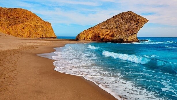 Ein Strand mit sandigem Ufer und einer großen Felsformation im Wasser.