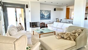 Ein modernes Wohnzimmer mit Sofa, Sessel und einem Glastisch. Ein Kamin, ein Gemälde an der Wand und ein Küchenbereich sind im Hintergrund zu sehen.
