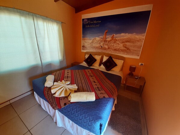 A bedroom with a bed, a nightstand, a wall-mounted lamp, a window with curtains, and a poster of a desert landscape.