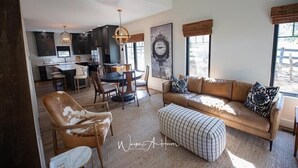A spacious living area with a leather sofa, a coffee table, and a dining table with chairs. There is a kitchen in the background with dark cabinets and a pendant light. - Discover Your Perfect Getaway: Cottage in the Heart of Horse & Bourbon Country! (Midway)