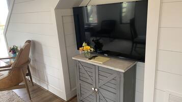 Une pièce comprenant un grand téléviseur à écran plat fixé au mur, un meuble gris avec des tiroirs, une chaise et une petite table sur laquelle est posé un vase de fleurs.