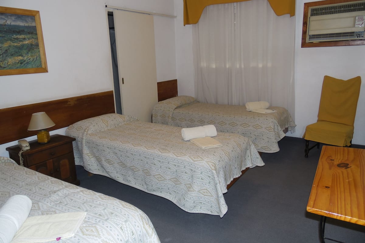 A hotel room with three beds, a wooden nightstand with a lamp, a chair, and a table.