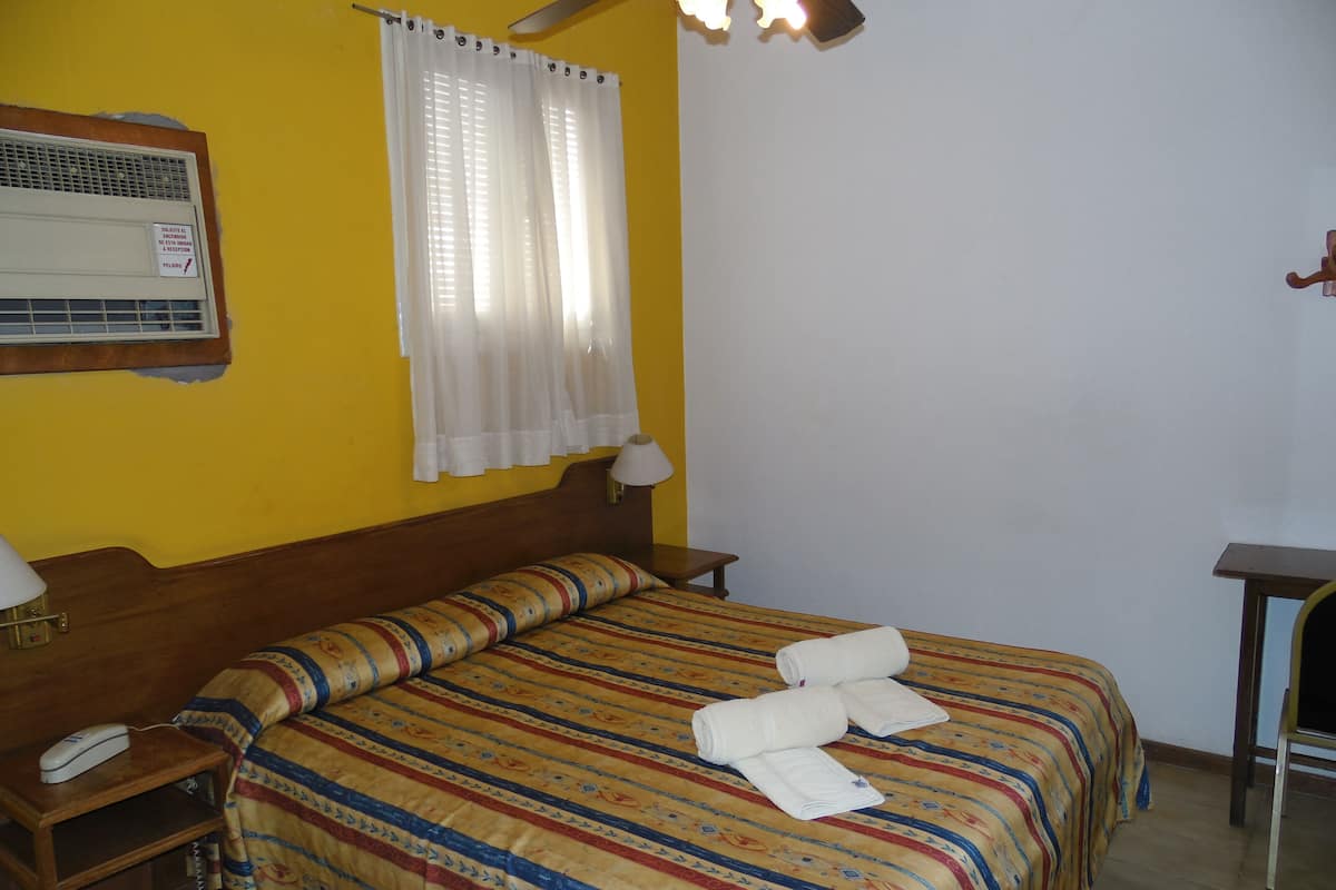 A hotel room with a bed, a bedside table with a lamp, a telephone, and a window with curtains.