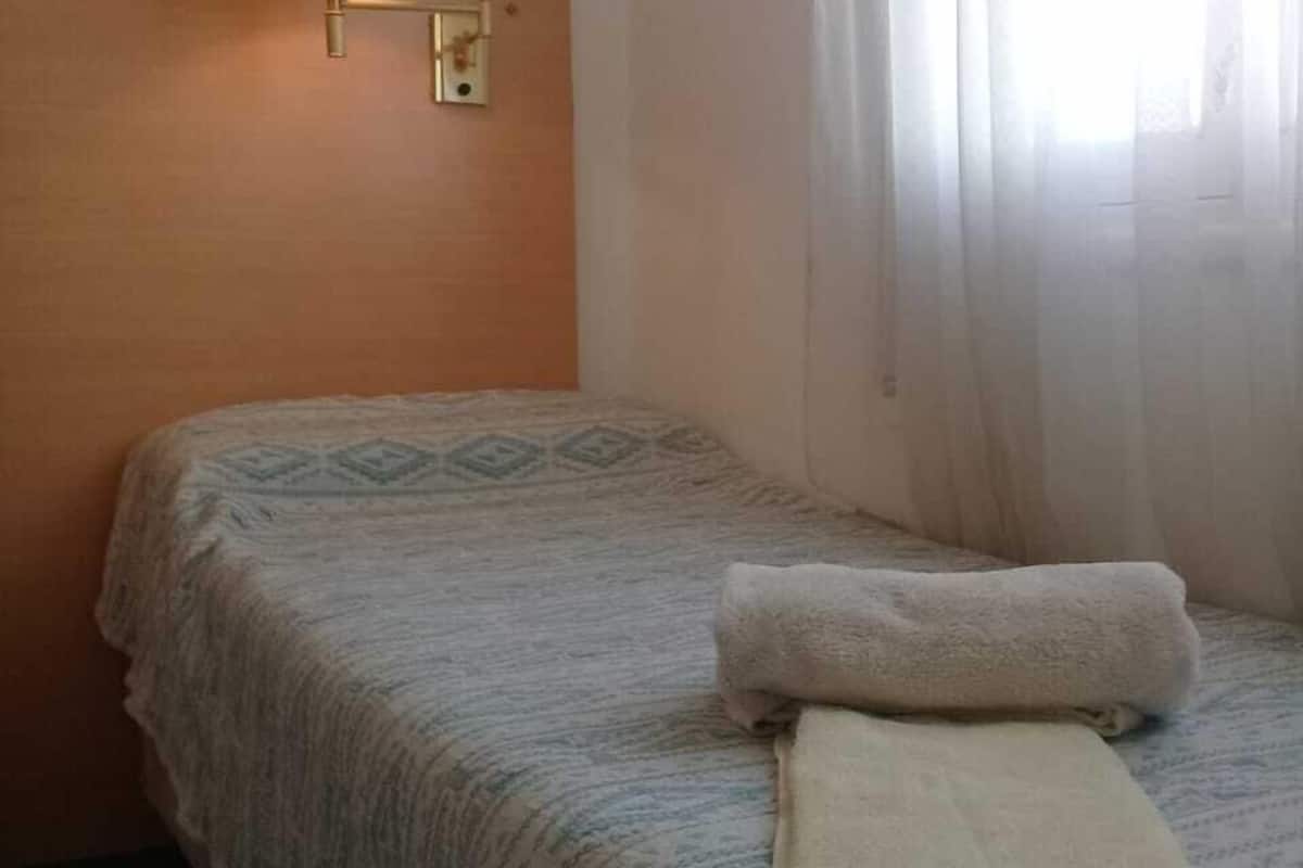 A single bed with a patterned bedspread and a folded towel, a wall-mounted lamp, and sheer curtains.