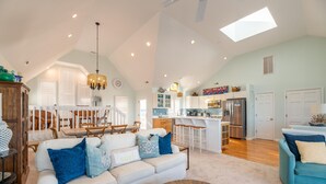 A spacious living area with a white sofa, blue cushions, and a coffee table. A kitchen with white cabinets and a stainless steel refrigerator is visible in the background. - Point of View - Nags Head MP 15, Oceanfront, Ocean View, Elevator, Pool, Hot Tub, Gazebo (Nags Head)