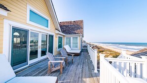 A wooden deck with outdoor seating, a view of the beach, and a house with large windows. - Point of View - Nags Head MP 15, Oceanfront, Ocean View, Elevator, Pool, Hot Tub, Gazebo (Nags Head)
