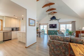 A spacious living area with a high ceiling, wooden beams, a ceiling fan, a large television, and a dining area with a view.