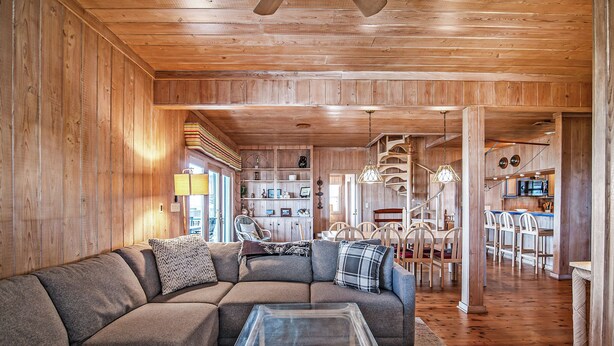 Un salon chaleureux avec un canapé d’angle, une table basse en verre, un plancher en bois et, au fond, un coin cuisine.