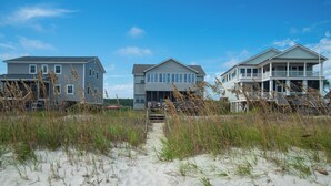 Une propriété située en bord de mer, comprenant trois maisons, un sentier sablonneux menant à la plage et de hautes herbes au premier plan.