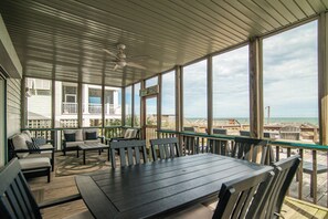 Ein überdachter Außenbereich mit Blick auf den Strand, ausgestattet mit einem Tisch, Stühlen und einem Deckenventilator. Ein Schild trägt die Aufschrift 'BEACH'.